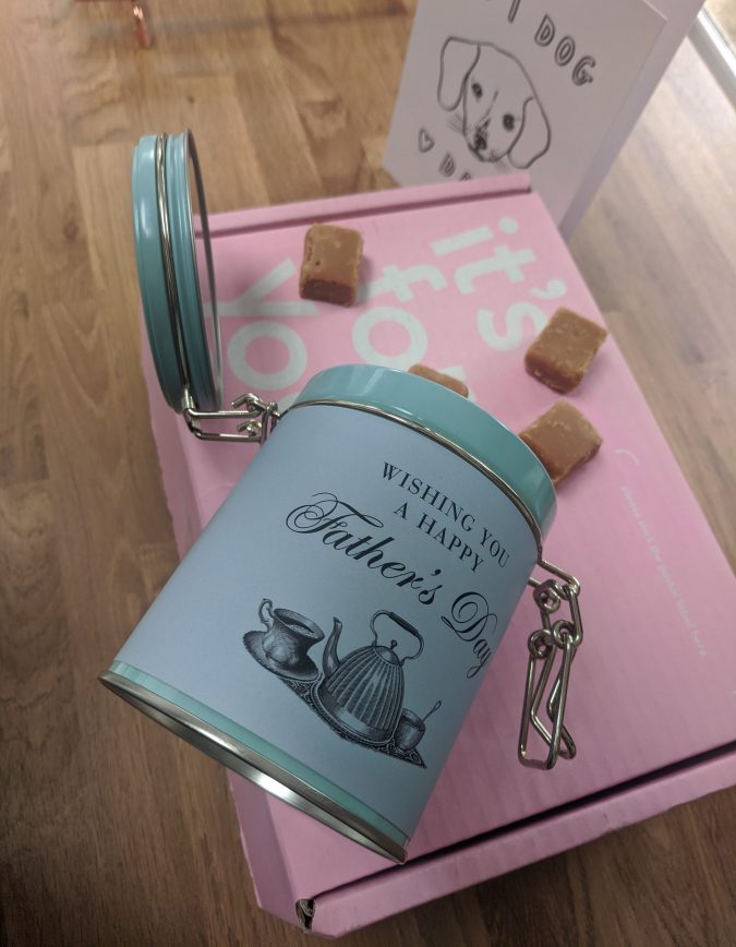 Father's Day 2020 How To Celebrate, Whether Near Or Far. Pink Moonpig Cardboard box, white father's day card with dog on it, blue tin of fudge with Happy Father's Day, with Salted Caramel Fudge spilling out
